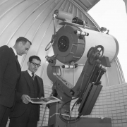 Two unidentified men look at a star chart in a book beside a telescope in an unidentified observatory, probably in Colorado. The telescope is mounted on a large hydraulic base and has a eye-piece. Lettering on a piece of equipment mounted on top of the telescope reads: "General Electric."
