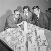 Three men look at a model of a proposed building in Denver, Colorado. The model encompasses several blocks of downtown Denver, one of the men has his hand on the top of the proposed building.