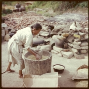 A Korean woman stands near a large wooden mortar filled with grain and grinds it with a stone pounding mallet with a wooden handle in South Korea. She wears a linen dress and her hair is pulled back in a bun. A flat winnowing basket and iron cooking pots are in the distance.