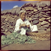 A elderly Korean woman sits near a thatched stone house and prepares greens in South Korea. She wears a linen dress and her hair is pulled back. A large basket and a pile of unidentified greens are beside her.