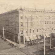 View of the corner, four-story Daniels & Fisher building at 16th (Sixteenth) and Lawrence Streets in the Central Business District in Denver, Colorado. Windows have hood molding and awnings and the flat roof line has decorative brackets and finials. Shows horse drawn carriages in the street, street car tracks on 16th Street and probably telephone poles. A sign reads: "W.F. Perkins, Cigars [and] Tobacco."