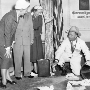 Colorado Governor Daniel Thornton kneels near a wastepaper basket on the floor of his office in the state capitol building in Denver, Colorado. He wears coveralls over his suit and lifts the edge of a mat. The floor is littered with paper and he holds a piece in his hand. He wears a cowboy hat and boots. Women and a man look on and laugh. A sign on the wall reads: "Governor Thornton Swept Here."