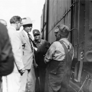 Colorado Governor Daniel Thornton stands and talks to two Denver and Rio Grande Western railroad employees near a railroad freight car in probably Denver, Colorado. He wears a suit, tie and a cowboy hat. The railroad employees wear overhauls and railroad caps. Members of the governor's staff are nearby.