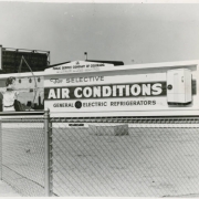 View of a billboard located in Denver, Colorado. The billboard has a frame is in an Art-Deco style with stylized newel posts on either side, molding and a cornice mounted on the top that reads: "Public Service Company of Colorado." A small plaque on the bottom of the sign reads: "Alden Sign Co."  The sign shows a woman in a shirt, pants and gloves, she aims a bow and arrow, the skyline of a city is behind her. Text reads: "For Selective Air Conditions" "General Electric Refrigerators."