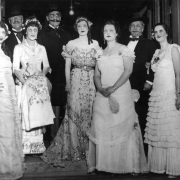 Men and women stand in a group on the steps of the Teller House in Central City, Colorado, in 1890's costume. The women wear long gowns of satin and lace with bustles. The men wear dark suits with string ties or ascots, wide-brimmed hats, and false facial hair. One of the men wears glasses.
