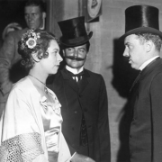 Men and a woman stand in front of the Teller House in Central City, Colorado; costume includes a satin dress, a shawl and flowers, suits, string ties, and top hats. The man in the center wears a false moustache. In the background, a man leans against the doorway of the Teller House and a Mountain States Telephone sign is on the exterior wall of the hotel.