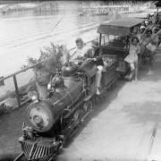 A group of women pose with the conductor on a miniature train at Lakeside Amusement Park in Denver, Colorado. Lake Rhoda and a roller coaster are nearby.