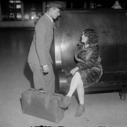 A man in a porter uniform talks to a woman seated on a wooden bench at Union Station, in Denver, Colorado. The woman wears a fur coat and has a suitcase.