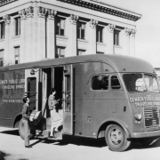 View of the Denver Public Library bookmobile in Denver, Colorado; lettering reads: "Denver Public Library - Traveling Branch." A man and woman board the vehicle; the Carnegie Building of the Denver Public Library in Civic Center Park is in the background.