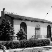 View of the Elyria branch of the Denver Public Library located on 47th Avenue and High Street in the Elyria-Swansea neighborhood of Denver, Colorado. The spanish style building was designed by architect H. J. Manning; it features a tile roof, arched windows, iron lamps, and a projecting doorway. The Elyria branch was closed in 1952.