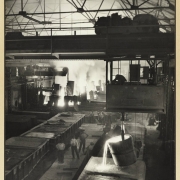 Interior view of the Colorado Fuel and Iron Company Minnequa Plant in Pueblo (Pueblo County), Colorado, shows machinery, scaffolding, and bucket pouring molten steel. Steam or smoke rises from machinery nearby, men watch from the walkway.