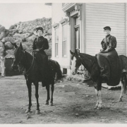Women sit sidesaddle on horses at 5th (Fifth) and High Streets in Central City (Gilpin County) Colorado; outfits include puff sleeved blouses. A house with volutes and pediments is by rocky crags.