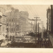 A view of Lawrence and 15th (Fifteenth) streets in Denver, Colorado; shows pedestrians, cable cars, utility poles, smokestacks, and commercial buildings. Signs read "Wright Clothing" and "Golding, Loftus & Breunert Carpets and Draperies.