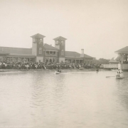 View of the pavilion near City Park (Ferril) Lake in City Park, Denver, Colorado. The Spanish Colonial Revival style building has towers with tile roofs and an arcade. People sit on chairs at the shore of the lake with parasols as groups in rowboats go by.  A band can be seen in the bandstand on the lake.