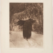 A Native American (Sioux) woman hauls a bundle of sticks on her back. The woman wears a woven, striped blanket over her shoulders and head, and walks on a snow covered hillside.
