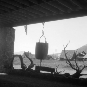 View from the Ore Bucket Lodge in Crested Butte, Colorado; shows the silhouettes of an ore bucket hanging from a rafter on a chain, and a large tree branch laying under the bucket; two large stone columns and a roof form a frame for the two objects; image taken from shadow into light; residences and mountains in the background.