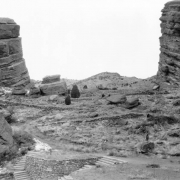 A woman in a sweater and a plaid skirt stands on a rock and looks at rock formations in Red Rocks Park, a Denver Mountain Park in Jefferson County, Colorado. Shows a dirt path, flagstone stairs and stone retaining walls.