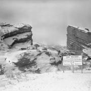 View of a sign near the unfinished Red Rocks Amphitheater in Red Rocks Park, a Denver Mountain Park, in Jefferson County, Colorado. The sign reads: "This Theater is Under Construction by the Civilian Conservation Corps Under Direction and Supervision of the National Park Service." Shows snow covered rock formations, concrete seats, and stone buildings.