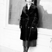 Grace Bedford poses in Keota, Weld County, Colorado, wearing  black shoes, stockings, skirt, and fur coat. She has black hair, cut in a "bob." Windows in a frame wall are behind her.