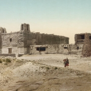 View of San Esteban del Rey de Acoma, a mission church, and the convento (convent) at Acoma Pueblo, New Mexico. The buildings are constructed of adobe and stone. The church has two bell towers; the two-story convent is attached to the mission building and has a balcony with a wooden railing and stone buttresses. Girls stand near the buildings.