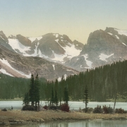 View of Long Lake and Navajo Peak, Apache Peak, and Shoshoni Peak, in the Indians Peaks in Boulder County, Colorado. A man stands on a spit with trees on the lake.