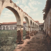 View of an arcade at Mission San Juan Capistrano in San Juan Capistrano, California. Plants grow from the top of a colonnade, and rubble is piled against a wall.  Plants grow wild in the central patio of the mission. The adobe walls and flagstone arches of the mission have missing plaster. The buildings have red clay tile roofs.