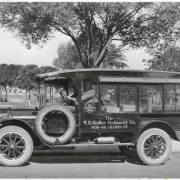 View of a Ford Model T delivery truck in City Park, Denver, Colorado. Lettering on the truck reads: "The Colorado House" and "The W.H. Kistler Stationery Co. 1636-46 Champa St." Two men sit in the truck. The bed of the truck has wire mesh and rolled-up canvas covers. Packages are in the back of the truck. A metal cylinder is mounted on the running board of the truck. Trees, sidewalks and people are in the distance.