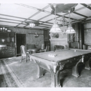 View the billiard room at Cleveholm (Osgood Castle), the home of John Cleveland Osgood, located in Redstone (Pitkin County), Colorado. The room has a large, carved billiard table, a fireplace flanked by two glass fronted cases, a carved sideboard and a table and chairs. A bronze chandelier with two glass shades hangs over the billiard table. An oriental rug is on the floor.