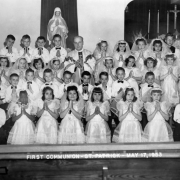 Holding prayer books with hands poised for prayer, This communion class (one Black student) in Holyoke, Phillips County, Colorado poses in white shirts and taffeta dresses. In vestments, a priest is in the  back row; behind them are an altar with candles and flowers, and a likeness of the Virgin Mary.