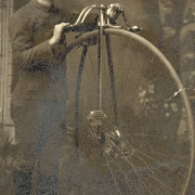 Charles A. Chase, probably a resident of Georgetown (Clear Creek County), Colorado, poses in front of painted backdrop with a high wheel bicycle. The young man wears a loose buttoned jacket with a pleated front, matching knickers, dark long stockings and shoes. He has a cap with a small bill set back on his head.