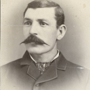 Studio portrait (profile) of Charlie Hurley, probably a journalist, an employee of the Colorado Miner newspaper, and resident of Georgetown (Clear Creek County), Colorado. He wears a coat buttoned at the top with a collar with bound edges and a patterned silk necktie.  His dark hair is parted on the side and he has a full, waxed mustache.