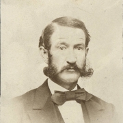 Studio portrait (profile) of John R. Hamble, an insurance agent and resident of Georgetown (Clear Creek County), Colorado. He wears a coat with large lapels, a vest and an oversized, silk bow tie.  His dark hair is parted on the side and he has sideburns and a long mustache curled at the ends.