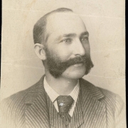 Studio portrait of Frank Graham, probably a boarding house keeper in Idaho Springs (Clear Creek County), Colorado. He wears a matching striped vest and coat with wide lapels, a checkered shirt, and a patterned, silk tie secured with a stick pin.  His dark hair is receding and parted on the side and he has wide sideburns that connect to a thick mustache (burnside).