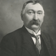 Studio portrait of Andrew W. Sylvester, a saloon keeper, miner and resident of Georgetown (Clear Creek County), Colorado. He wears a dark wool jacket, a shirt with an upstanding, wing collar and a striped tie with a stick pin. He is balding, his hair is parted and combed into a cowlick, and he has a walrus mustache.