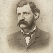 Studio portrait of Edward C. Parmlee, postmaster and resident of Georgetown (Clear Creek County), Colorado. He wears a striped, wool, sack coat buttoned at the top, a shirt with an upright, wing collar and a bow tie. His dark, curly hair is parted on the side and he has a full mustache and bushy sideburns.