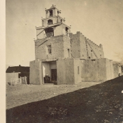 Men pose in the entrance or narthex of the adobe Church of San Miguel in Santa Fe (Santa Fe County), New Mexico. One man, a priest, wears a cassock. The church has tapered towers with wooden railings and battlements along the roof line of the nave. Shows wooden lintels above the entrance and a wooden ladder at the side of the church.