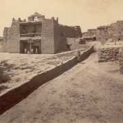 View of an adobe Nuestra Señora de Guadalupe mission church at Zuni Pueblo, New Mexico. Native American (Zuni) men pose in the entrance and on a second floor balcony. The church has two bells in a central belfry, worn battlements on towers, and vigas near the roof line of the nave. A cross and altar are in the walled courtyard. A man, probably a member of the survey crew, leans against an adobe wall.