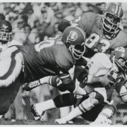 Two Denver Broncos professional football players tackle an opponent. Spectators are in the background.