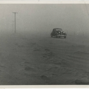 A two-door sedan approaches amid blowing dust. Telephone poles are to the sides.