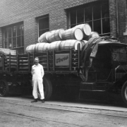 Len Vogel stands in his white overalls in front of his Coors delivery truck at the Adolph Coors Brewery, Golden, Jefferson County, Colorado. The truck has an open frame bed loaded with wooden barrels and another trailer attached with wooden crates. A canvas covering for the load  is on top the truck cab.