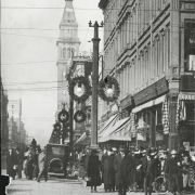 Pedestrians walk along bustling Sixteenth Street around noon at the intersection of Stout Street in the City and County of Denver, Colorado.  Christmas wreaths hang from the lightposts.  Cars and bicycles are parked along the street.  Landmarks include the Daniels and Fisher Tower.