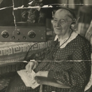Katherine Steiger smiles and wears headphones; the elderly woman wears a polka-dot dress with a lace collar, glasses, and holds a newspaper: "The Enquirer - Radio Section." A radio receiver is on a table; lettering between dials reads: "Crosley Trirdyn 3R3." A patterned, fringed cloth and antimacassar complete the interior.