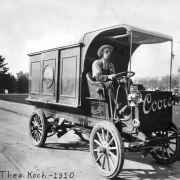 Theo Koch drives an Adolph Coors Golden Brewery delivery truck, in Golden, Jefferson County. The wood panel Frayer - Miller truck has the steering wheel on the right side, narrow tires and lantern headlights.