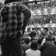 Students crowd the interior of the administration building at the University of Colorado at Boulder during a war protest. Seated people on second and third floor mezzanines let their lower legs dangle through the vertical bars. Men at the center read a paper, and a man in the foreground has a clenched fist stenciled on his flannel shirt.