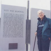 Bill Hosokawa, using a cane, standing in front of a panel at the Nisei War Memorial, located in Fairmount Cemetery in Denver, Colorado