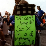 Photo of a handwritten sign taped to the back of a protester's wheelchair at the ADAPT protest against the American Health Care Association's 1994 Las Vegas convention.  Photographer unknown.