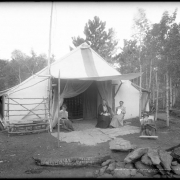View of a campsite in the Rocky Mountains of Colorado; large canvas tent is supported by wooden posts with awning extended over front door to make a "covered porch;" three women sit underneath awning (one weaves reed or cane, one reads, one needlepoints); a man sits at edge of "porch" reading McClure's Magazine dated September, 1906; shows blankets on  ground for carpet, mosquito netting over tent doorway, and split rail or log bench; a burned log and rocks form firepit ring, center foreground.