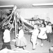 Students in fancy dress perform a maypole dance for assembled parents and teachers, inside a George Washington Carver Day Nursery schoolroom.