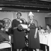 A member of the Negro Woman's Club Association presents a plaque for meritorious service to member Lillian S. Bondurant at an award ceremony. Audience members sit nearby. A sign for the United Way is on the stage behind the two women.