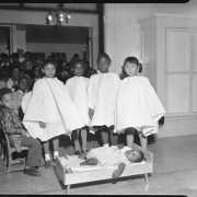Four girls in costume for a performance stand in a classroom of the original Carver Day Nursery building at 2357 Clarkson. The girls are arranged behind a small bed that holds a baby laying down. Additional boys and girls watch from the background.